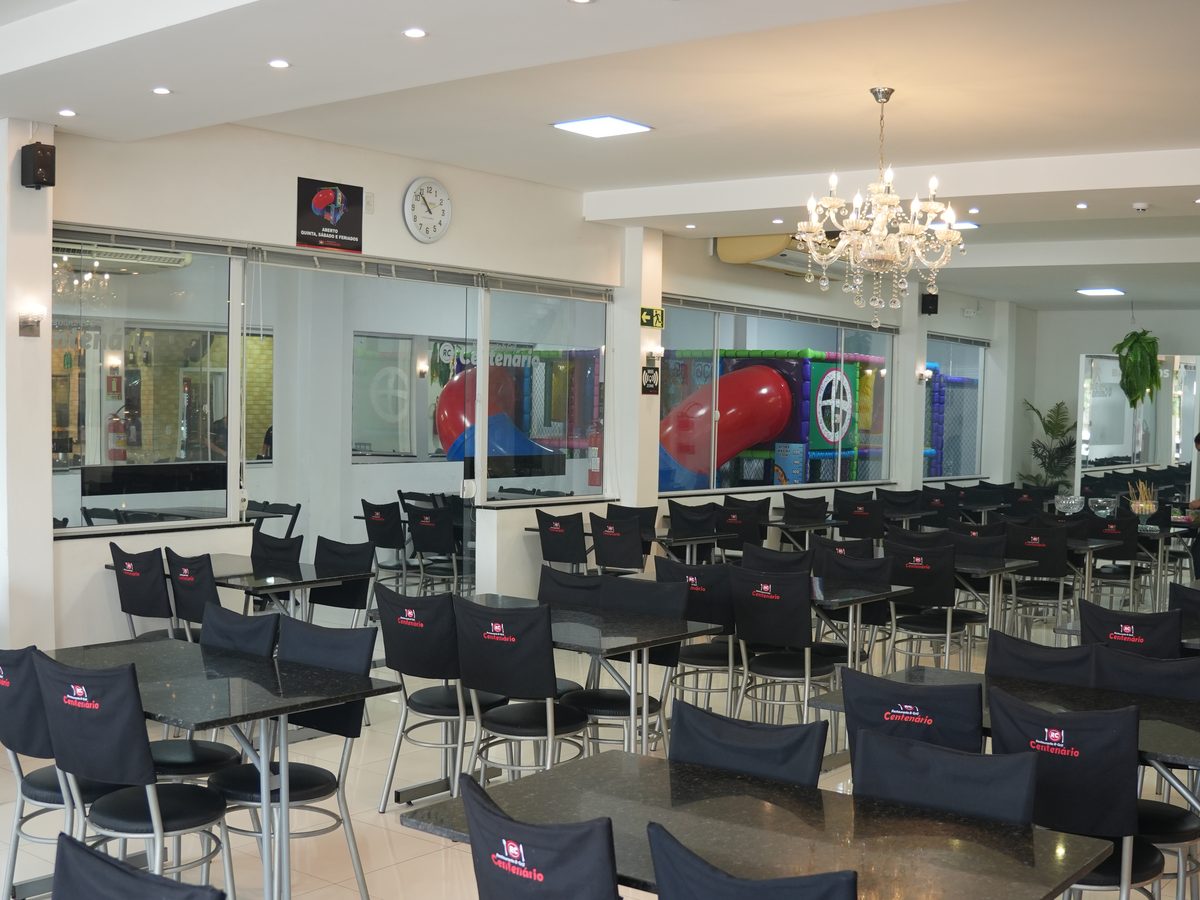 Salão do Centenário com mesas e cadeiras com a marca da casa; ao fundo, atrás do vidro, o espaço Kids.